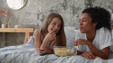 Two-female-friends-watching-TV-at-home.-Black-girl-and-caucasian-young-woman-eating-popcorn-lying-on-the-bed.