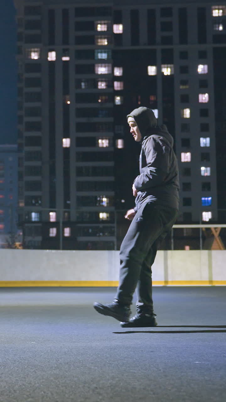 Side view of man in hoodie kicking soccer ball under bright stadium lighting at night, near goalpost and reflecting on wall. Urban cityscape with illuminated office buildings
