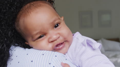 loving african american mother wearing pyjamas cuddling baby daughter in bedroom at home