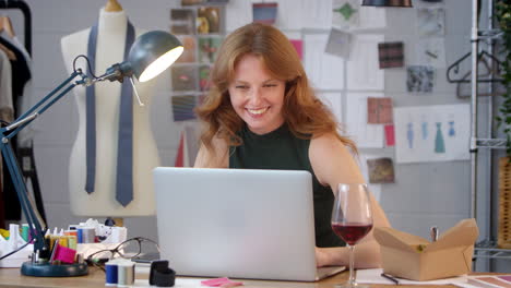 female owner of fashion business with glass of wine listening to music on laptop in studio