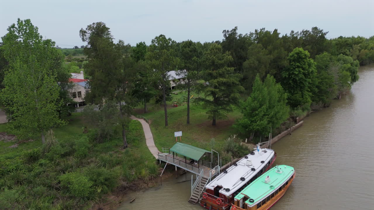 Slow drone panning clip next to river, parked boats and large house - building, with cloudy sky and vast vegetation surrounding.