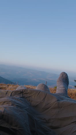 Romantic-tourist-couple-lie-in-tent-on-mountain-hill,-clink-cups-and-drink-tea.-Two-travelers-rest-under-blanket-during-adventure-vacation.-Hiker-family-warm-up-at-cold-weather-and-admire-the-scenery.