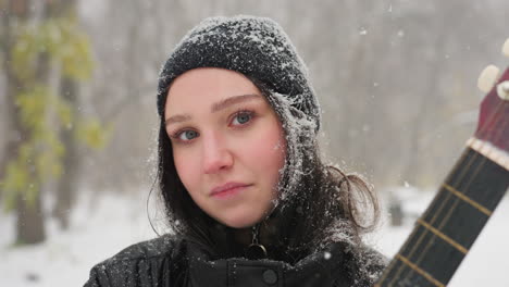 young girl standing holding a guitar in winter scene, snowflakes resting on her black beanie and jacket, staring directly at the camera with a calm and reflective expression