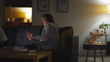 woman sitting on sofa at home at night streaming or watching movie or show on laptop and scrolling internet on phone 14