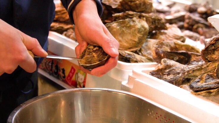 A vendor uses a knife to shuck fresh oysters at a busy outdoor seafood market.