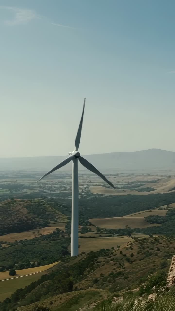 Video verticale: camera con panoramica che rivela una valle, alta turbina eolica che ruota sopra una cresta rocciosa
