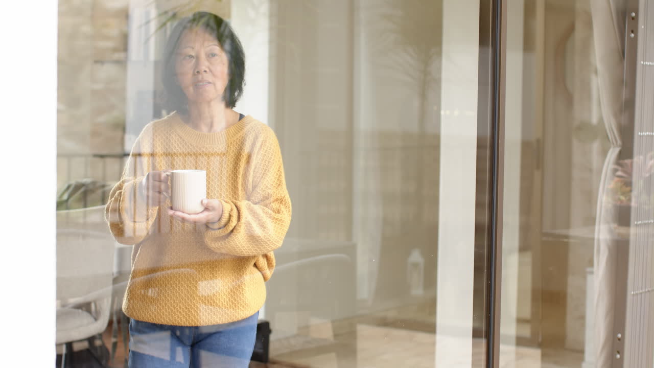 Senior Asian woman holding coffee mug, gazing thoughtfully out window at home, copy space
