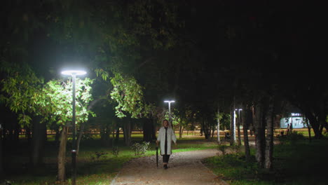 young girl dressed in white coat walking alone on peaceful park path at night illuminated by glowing streetlights, surrounded by trees, with blurred passing cars and people moving in background