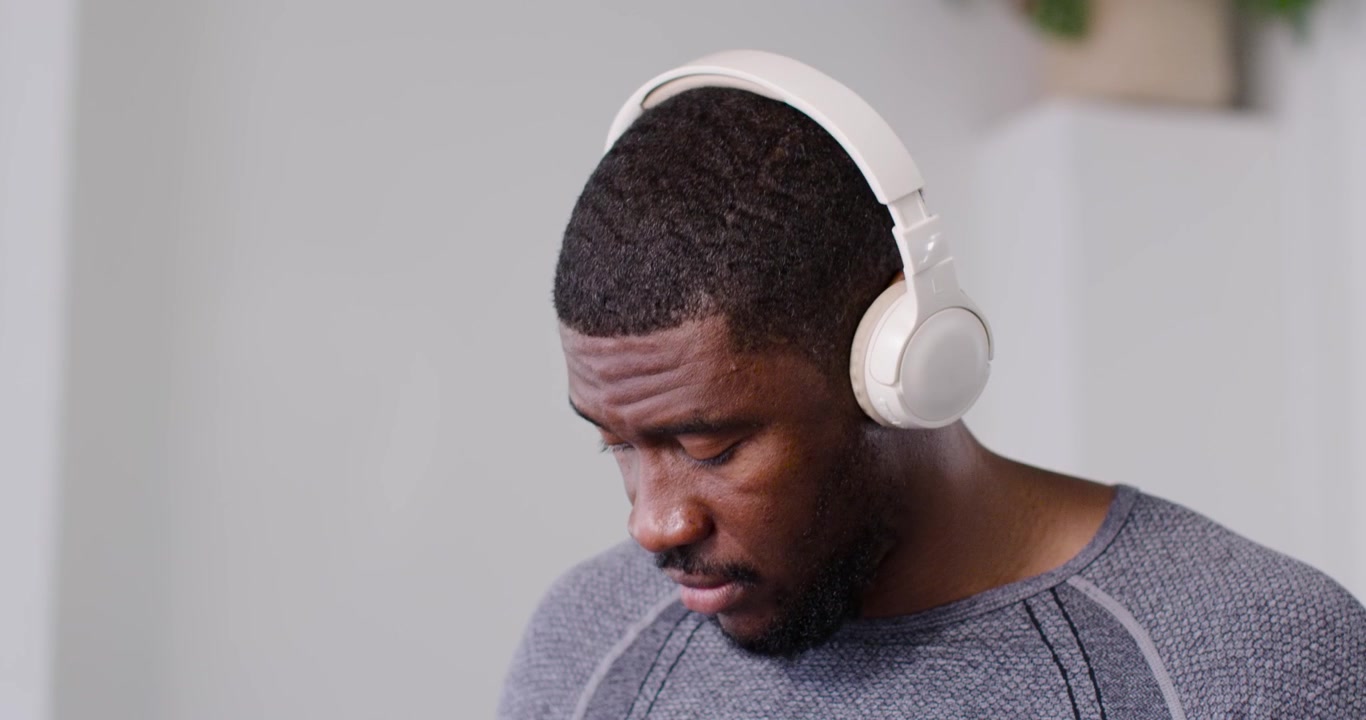 African man in gray top picking up, inspecting white headphones in gym, adjusting fit for workout