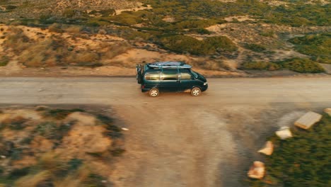 Viejo-Y-Nostálgico-Autobús-Furgoneta-Con-Tablas-De-Surf-En-El-Techo-Conduce-A-Lo-Largo-De-Una-Carretera-Polvorienta-En-Un-Paisaje-Mediterráneo,-Seguimiento-De-Drones