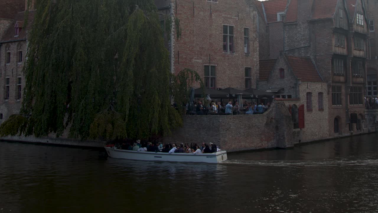 Tour boat glides past historic brick buildings and willow tree on tranquil Bruges canal