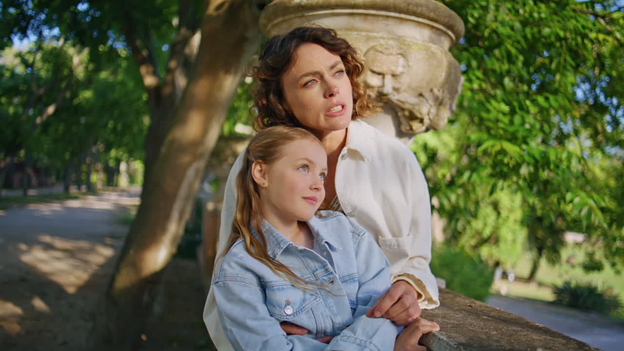 Caring mother bonding daughter pointing finger at sunny park. Family embracing