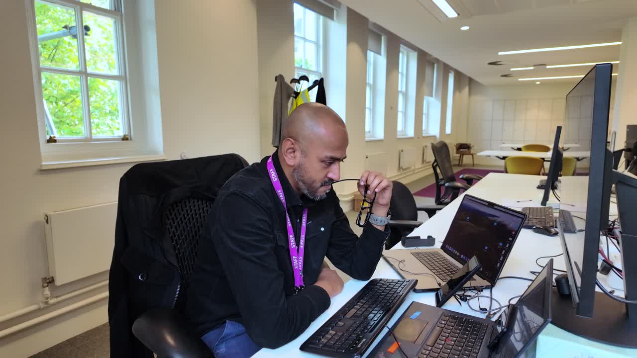 un individuo si siede a una scrivania in un ambiente di ufficio moderno, con gli occhiali in bocca e scuote la testa in frustrazione