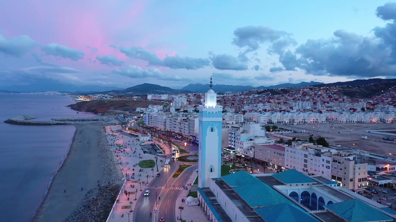 Veduta aerea all'ora blu della moschea sul lungomare di M'diq, del lungomare e della densa città costiera sotto i Monti del Rif sulla costa mediterranea del Marocco, Nord Africa