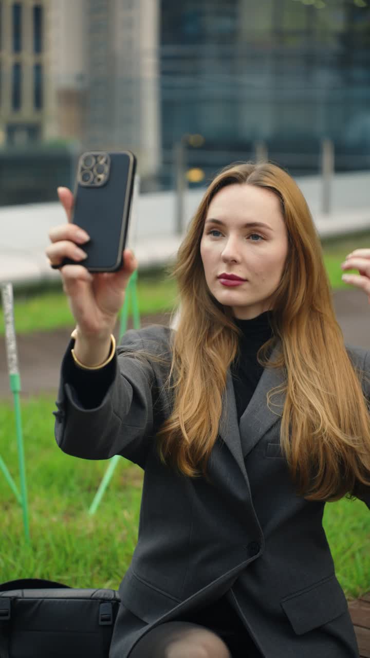 A stylish Caucasian woman styles her hair as she prepares to take a selfie with her phone on a modern terrace in Pangyo at dusk - slow motion
