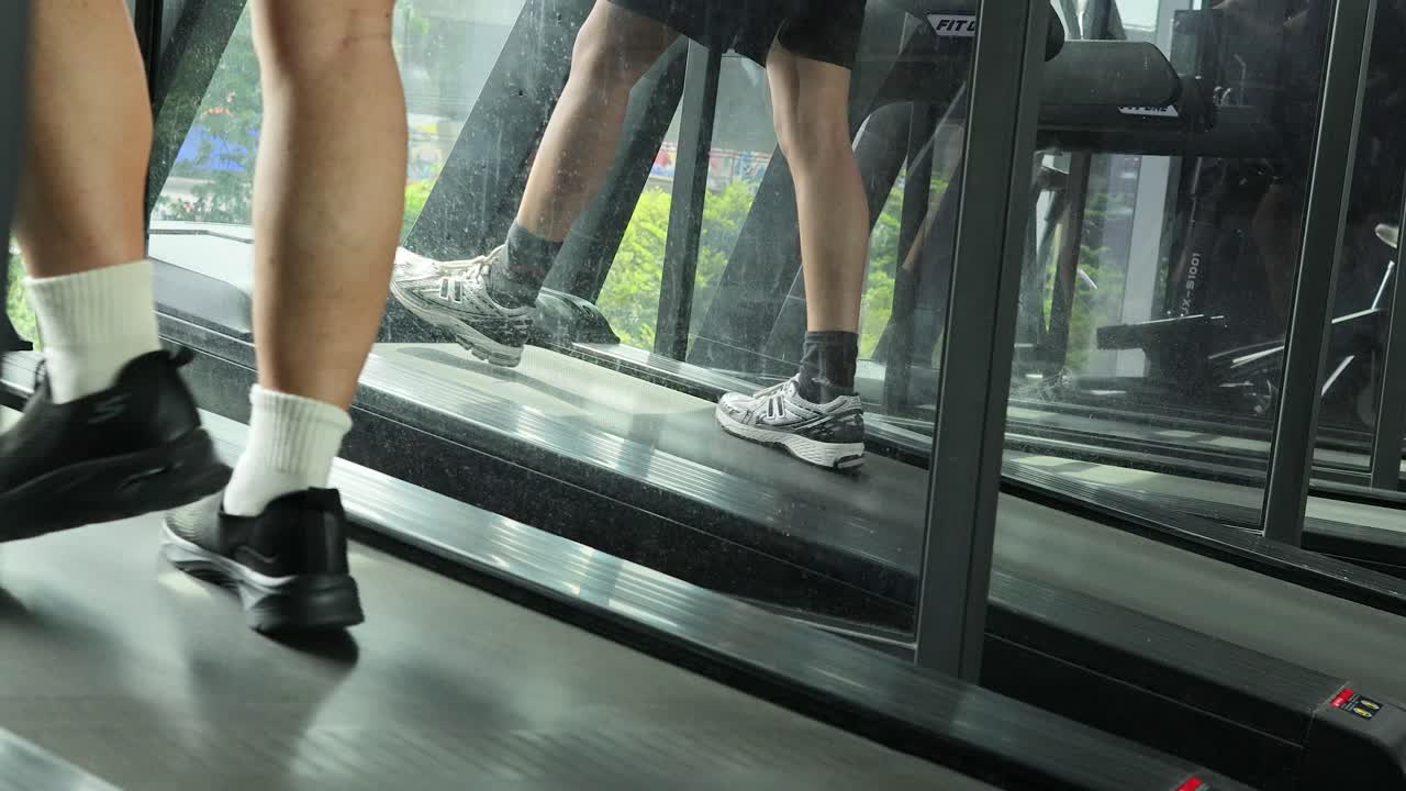 Close-up side view of a man’s legs running on a treadmill in a well-lit gym, with reflections and steady camera movement capturing dynamic motion