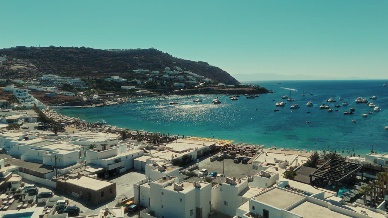 Drone ascending and moving sideways above Mykonos Bay, showing white houses, blue sea, and boats under sunlight reflection