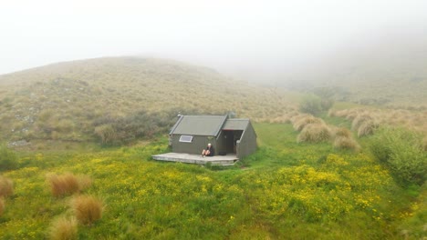 person sitting on small deck in front of hiker's hut within a flower field in the new zealand wilderness