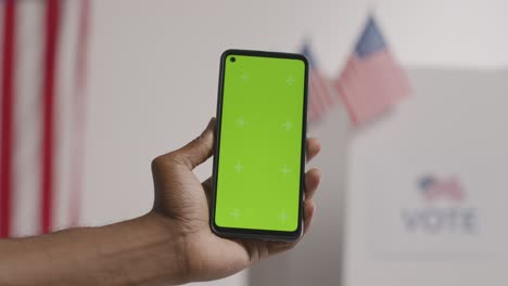 Close-Up-Of-Hand-Holding-Green-Screen-Mobile-Phone-In-Front-Of-Ballot-Box-In-American-Election