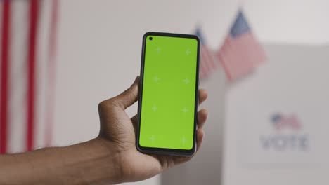 close up of hand holding green screen mobile phone in front of ballot box in american election