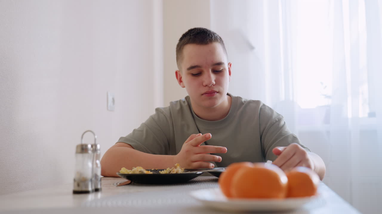 Ragazzino con maglietta verde si lecca le dita mentre è seduto a tavola, poi prende le posate per mangiare un piatto di uova strapazzate accanto alle arance in una stanza luminosa e soleggiata con interni semplici e un'atmosfera accogliente all'ora dei pasti