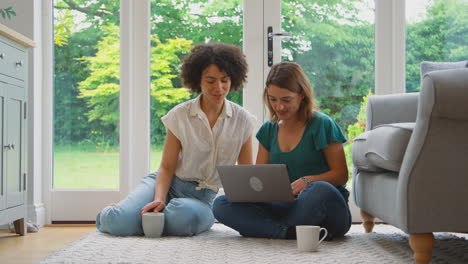 Same-Sex-Female-Couple-Or-Friends-At-Home-Sitting-In-Lounge-Using-Laptop-And-Drinking-Coffee