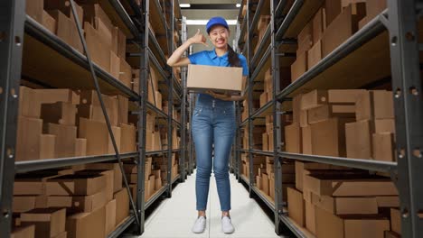 cuerpo lleno de mensajero femenino asiático en uniforme azul mostrando llámame gesto y sonriendo mientras entrega una caja en el almacén