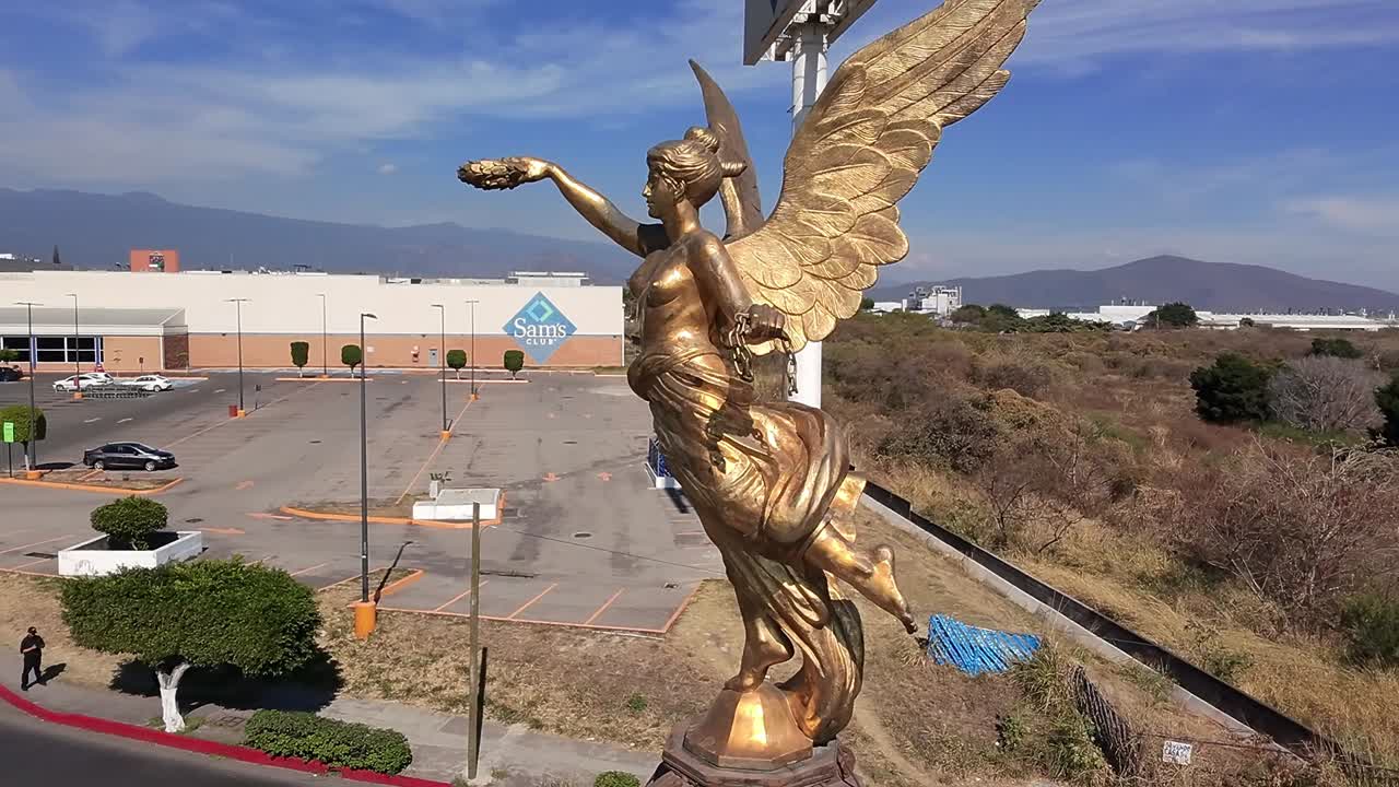 Golden statue of the Winged Victory holding laurel wreaths, located in Jiutepec, Morelos, Mexico, with a cityscape and mountains in the background during a sunny day