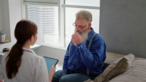 an elderly serbian man with hair and a white lush beard in glasses and a blue shirt communicates with a brunette girl doctor in a medical gown who is holding a tablet in her hands and recording the patient’s complaints during a home examination