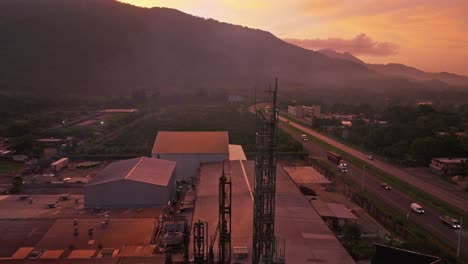 Toma-Aérea-Ascendente-De-La-Fábrica-Industrial-Y-El-Tráfico-En-La-Carretera-Durante-El-Amanecer-Dorado-Detrás-De-Las-Montañas---Villa-Altagracia,-República-Dominicana