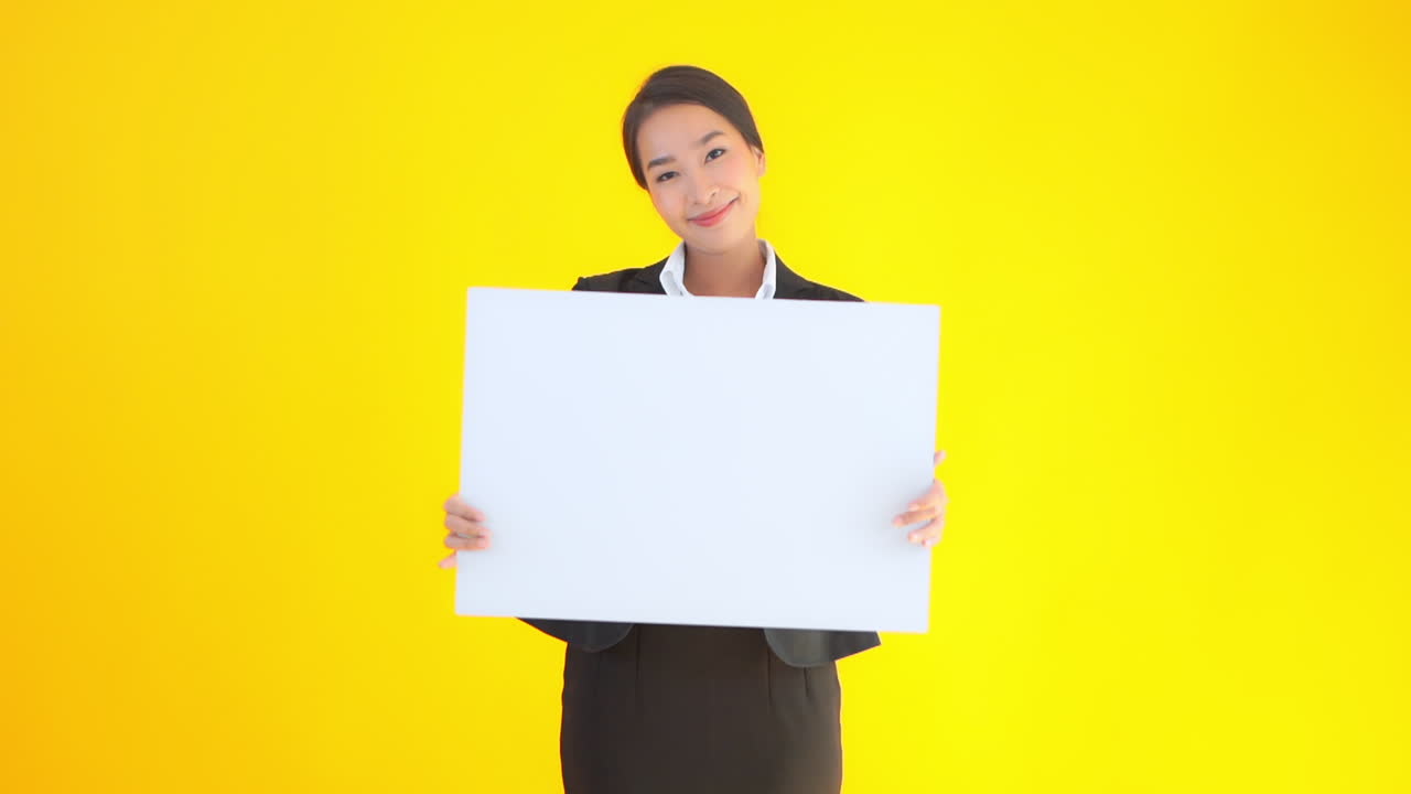 Asian businesswoman holding a blank sheet of the paper poster banner with a happy face expression on yellow studio background