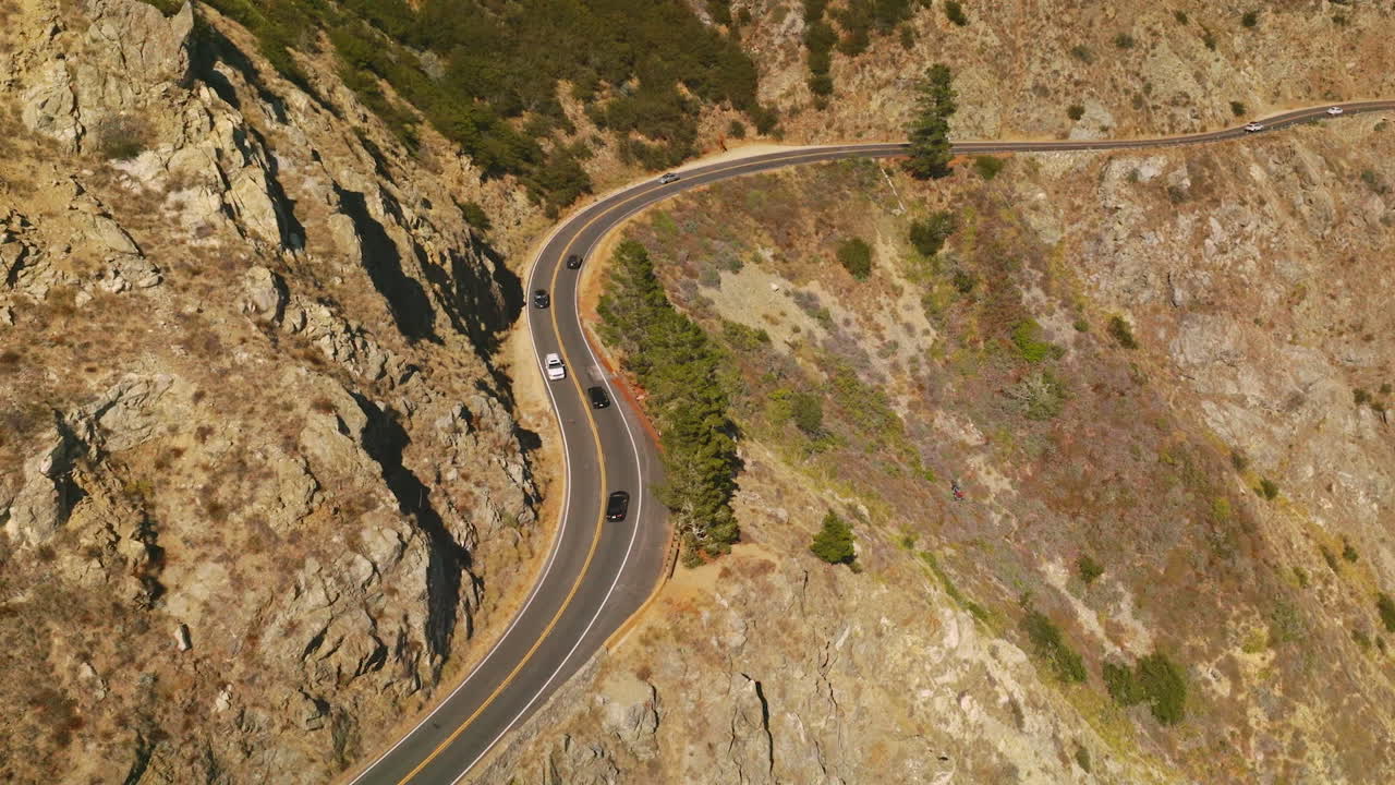 Lively road with cars moving back and forth in the mountainous landscape. Huge mountain with little greenery on sunny day from aerial view.