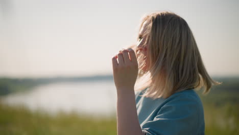 a close-up of a woman in a blue dress gently flicking and touching her hair as it flows in the breeze. she stands in a serene, blurred grassy field beside a lake