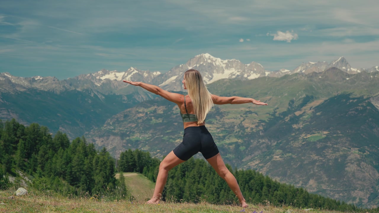 Atletisk blond kaukasisk kvinde udfører yogastillingen Krigeren på en alpeeng med udsigt over spektakulære sneklædte bjerge og Mont Blanc under en klar blå sommerhimmel.