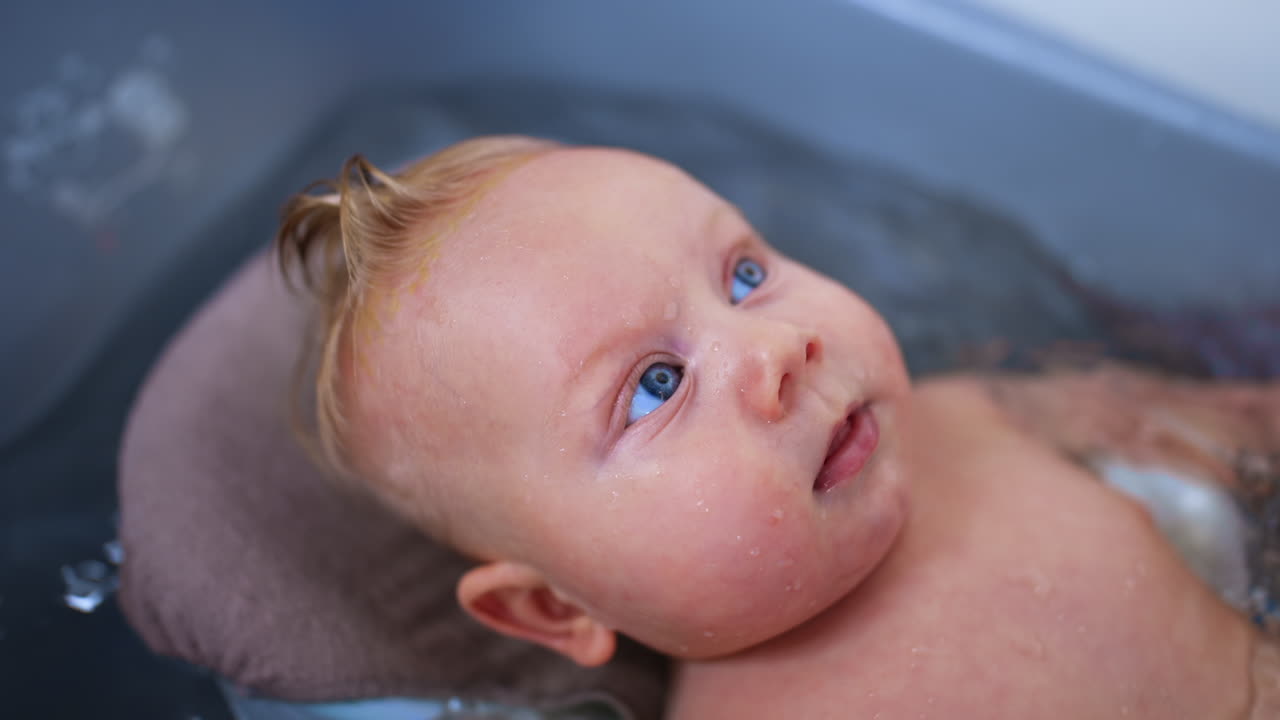 Caucasian baby with big blue eyes moving in the water. Cute child having a bath. Close up.