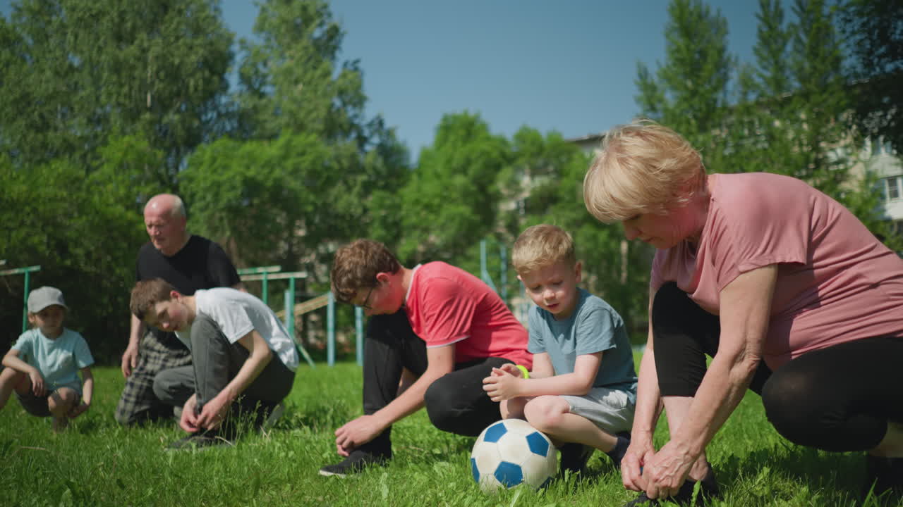 A warm family moment where a grandparent and their grandsons tie their shoelaces on a grassy field, the younger boy kneels on a soccer ball, looking towards his grandma