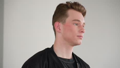 white boy wearing black jacket walks toward window with soft natural light filtering through, face illuminated by daylight, neutral walls and minimal interior