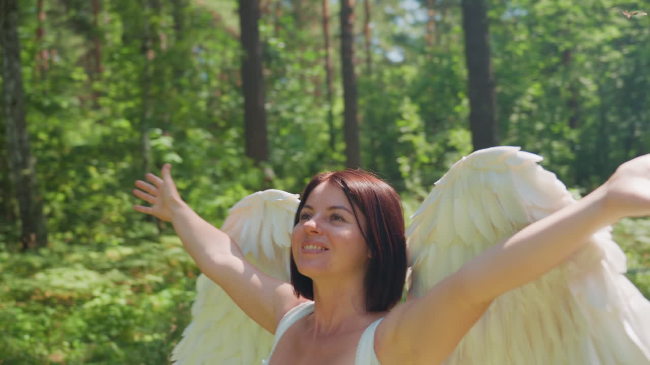 Close up of joyful girl with angel wings turning around under sunlight in lush forest, smiling warmly with eyes closed, expressing happiness, freedom, and peace surrounded by glowing leaves