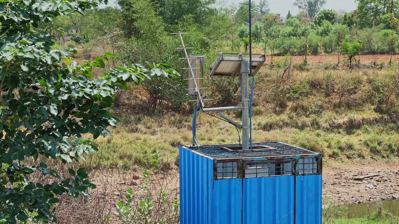 Piccola stazione di monitoraggio blu alimentata a energia solare con antenna e pannello solare installata vicino a uno specchio d'acqua rurale circondata da vegetazione secca!