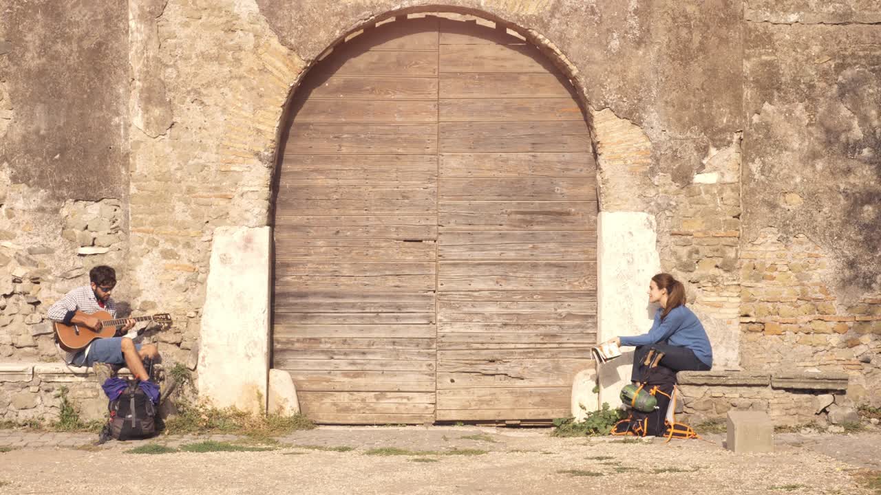 행복한 젊은 부부, 배 여행자, 기타를 연주하고 노래하는 관광객, 중세 건물의 목조 문 근처에 앉아, parco degli acquedotti에, 로마의 공원 폐허, 해가 뜨고, 지도 가이드, 느린 동작 스테이디캠.