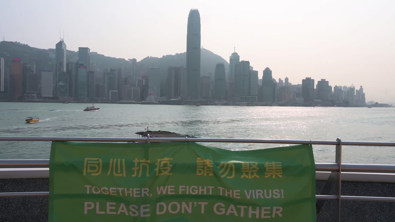 vista del horizonte de la isla de hong kong desde el paseo marítimo del puerto de victoria mientras una pancarta recuerda al público que evite las reuniones para prevenir la propagación del coronavirus en hong kong