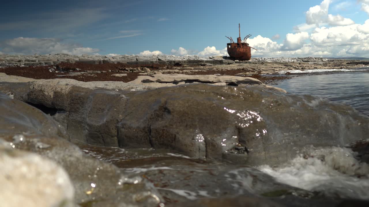 Ampia inquadratura cinematografica dell'iconico relitto della MV Plassy sulle coste rocciose di Inisheer, Isole Aran. Le onde dell'oceano si infrangono sulle rocce in primo piano sotto un cielo azzurro brillante