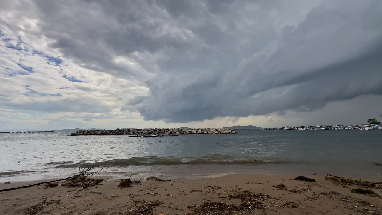 Dramatic dark storm clouds gather over a rough sea. Waves crash on the shore near a stone breakwater and a marina with boats on a gloomy autumn day
