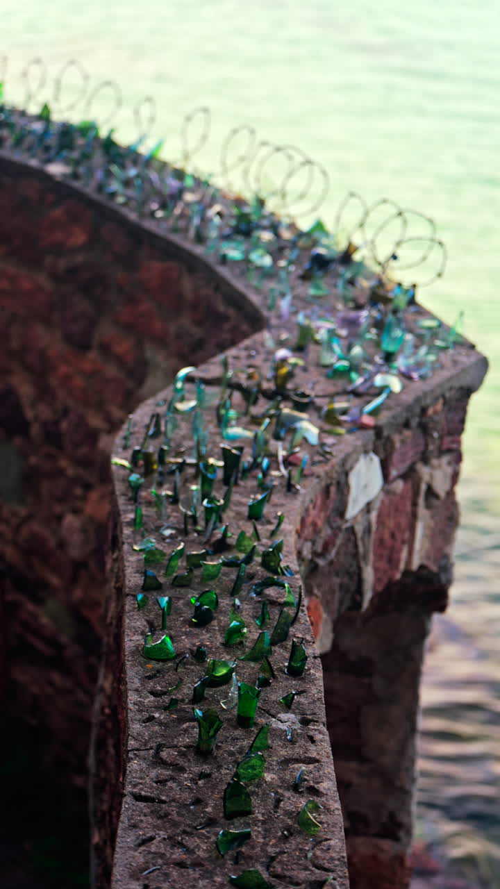 Close up of multiple broken glass shards and barbed wire loops along the edge of a brick wall with a blurred view of the sea. Vertical