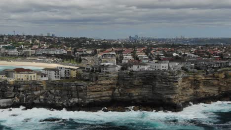 Waves-crashing-on-rocky-cliff-of-Ben-Buckler-Point-with-Bondi-Beach-in-background,-Australia