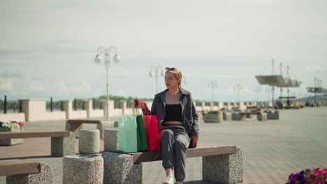 lady sits down on outdoor bench, drops her shopping bags, and adjusts red shopping bag, she looks to the right with a relaxed expression, surrounded by park benches, flowers, and street lamps