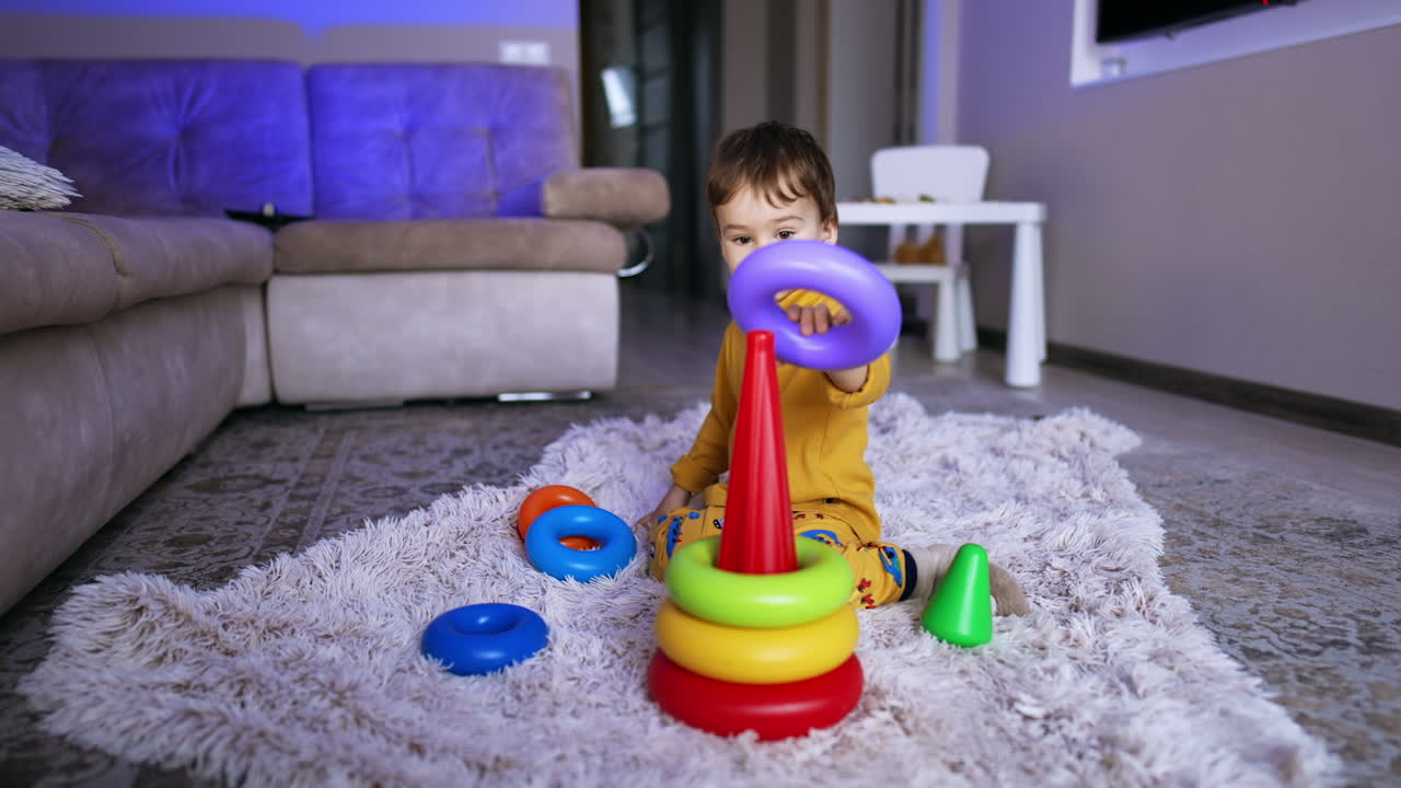 Peaceful Caucasian kid sitting on soft plaid on the floor. Cute toddler assembling pyramid.
