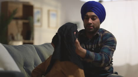 Father-Tying-Turban-Onto-Head-Of-Young-Sikh-Son-With-Top-Knot-Sitting-On-Sofa-At-Home-Shot-In-Real-Time