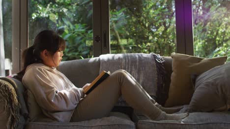 asian girl smiling while reading a book while siting on the couch at home