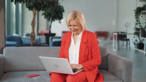 retrato de una mujer mayor con un traje de negocios rojo, sentada en un sofá en un centro de negocios con una computadora portátil, hablando y enviando mensajes en línea con una sonrisa en su rostro